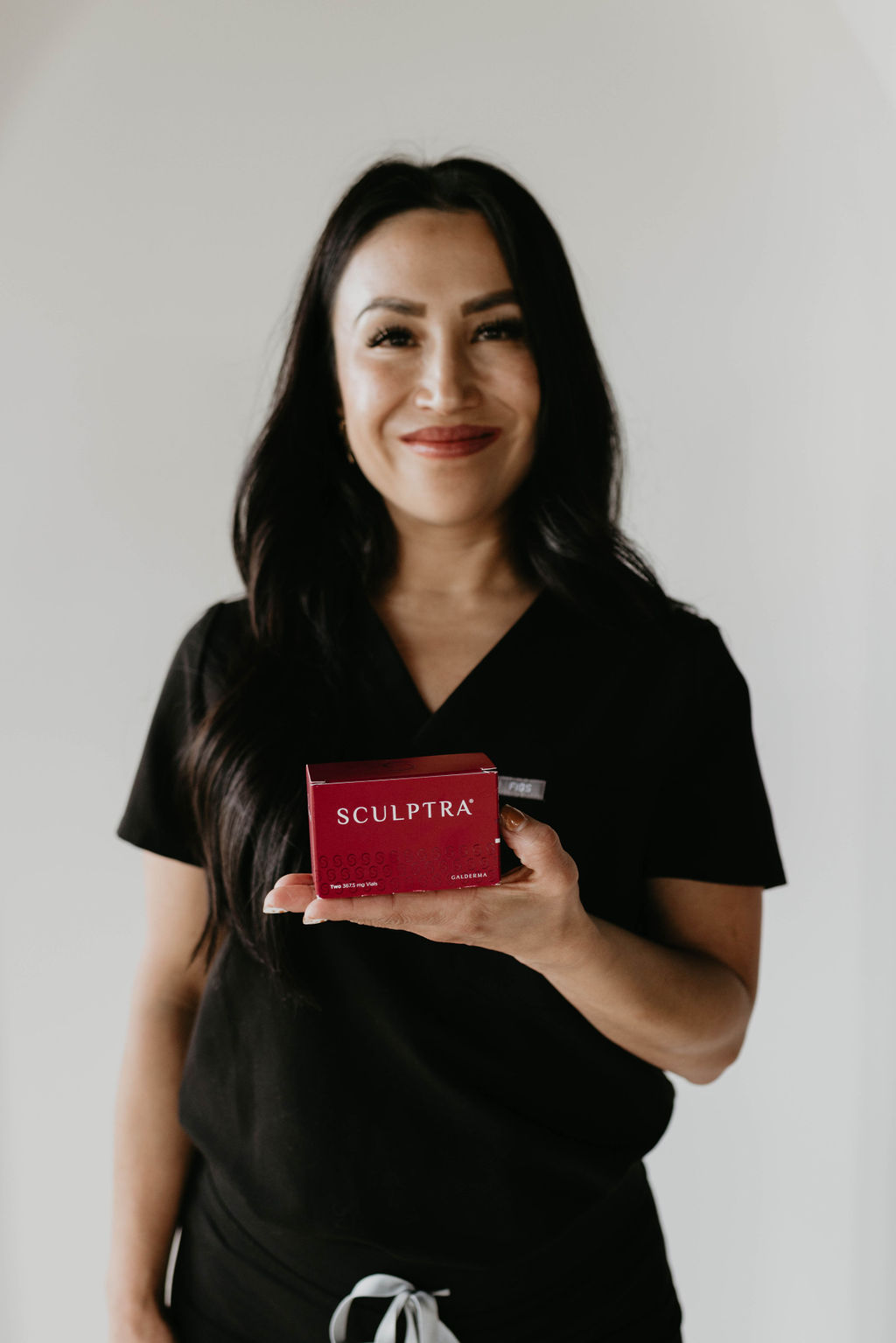A nurse injector in black attire holding a red product Sculptra box, smiling and standing in a professional setting.